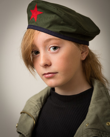 Studio portrait of a teenage ginger girl with long hair wearing a Che revolution barret hat and a green army jacket isolated against a grey background.の写真素材
