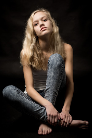 Studio portrait of a beautiful blond teenage girl with long hair wearing a white top and blue jeans sitting on the floor isolated against a black background.の写真素材