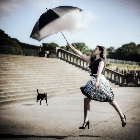 Artistic grunge style composite photo of a woman with black top, dotted skirt and long hair jumping up and catching a dream cloud with her umbrella in front of concrete stairs and a dog in the background.の写真素材