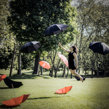 Grunge style photo of a woman with long black hair and wearing a black dress flying through the air and forrest holding an umbrella surrounded by more umbrellas.の写真素材