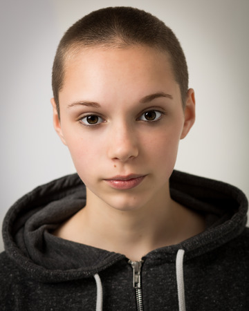 Studio portrait of a beautiful young teenage girl with a shaven bald head wearing a grey hoodie isolated against a light grey background.の写真素材
