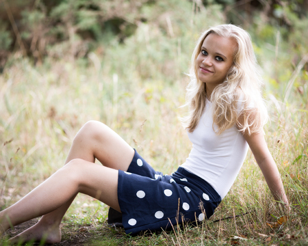 Portrait of a beautiful teenage blond girl with long hair wearing a tight white shirt and a blue skirt outside in a natural wood.の写真素材