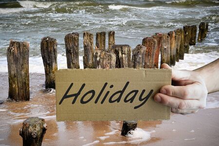 A hand holding a sign that says "Holiday" on the Baltic Sea beachの写真素材