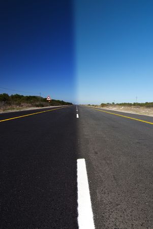 An road with clear blue sky above. One half is in full daylight, and the other half in darknessの写真素材