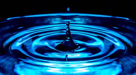 Flying drop of water macro with rippling water surface in neon blue colorsの写真素材