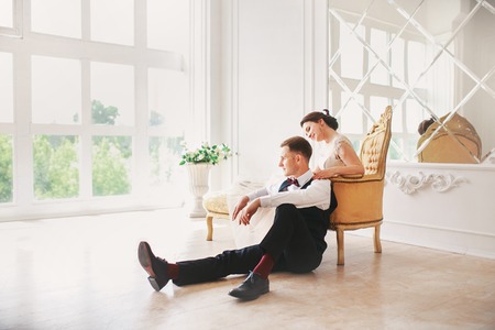 Bride in beautiful dress and groom in black suit sitting on sofa indoors in white studio interior like at home. Trendy wedding style .bride huggging Groomの写真素材