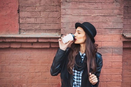 young beautiful brunette drinking coffee walking around the city.leather jacket,urban backpack , bright red lips and hatの写真素材