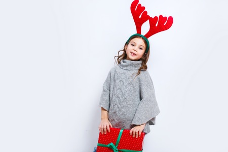 Happy little smiling girl with christmas gift box looking in camera. Christmas concept. Smiling funny girl in deer horns in studio.の写真素材