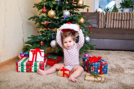 Happy cute baby girl in Santa Claus hat holding Christmas gifts at christmas tree at homeの写真素材