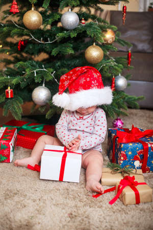 Happy cute baby girl in Santa Claus hat holding Christmas gifts at christmas tree at homeの写真素材