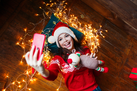 Close-up portrait of woman in Santa hat wrapped in christmas lights taking selfie and lying on wooden floor at homeの写真素材