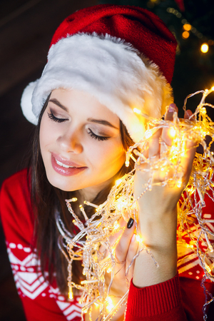 Beautiful young woman in Santa hat smiling and talking garlands of lights at christmas background with boke lightsの写真素材