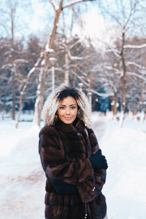 Outdoor close up portrait of young beautiful fashionable woman with afro hair , wearing fur coat in winter park. Winter fashion, Christmas holidays concept.の写真素材