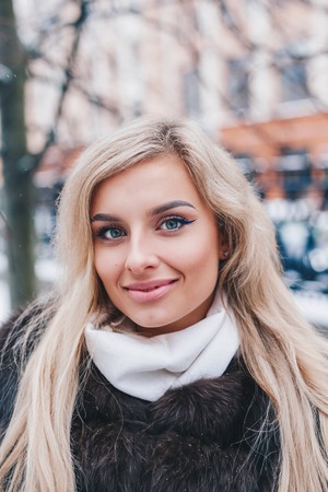 Outdoor portrait of young beautiful fashionable happy smiling girl wearing trendy fur winter coat,, posing in snow covered street.の写真素材
