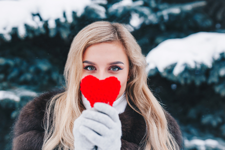 Portrait of a beautifull smiling woman in winter forest holding red Valentines heart in the hands.の写真素材