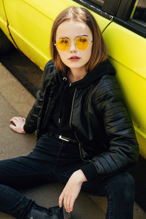 Beautiful fashionable teenager girl in sunglasses and leather jacket posing over yellow car outdoorの写真素材