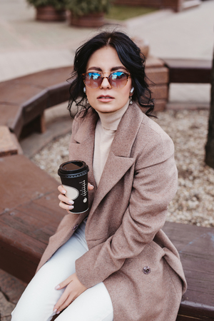 Beautiful elegant woman in coat and sunglasses sitting on the bench and drinking coffee outdoorの写真素材