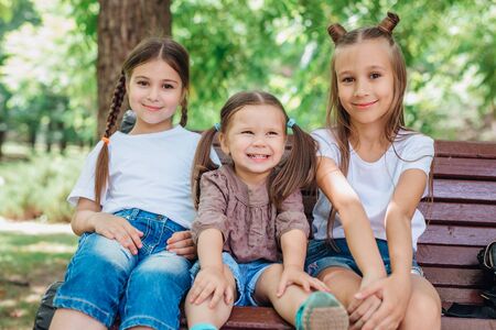 Three cute little sisters smiling and sitting on the wooden bench in summer parkの写真素材