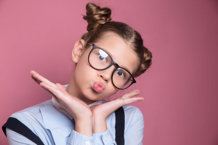 Close-up portrait of a young school girl with glasses having fun and looking at camera on pink backgroundの写真素材