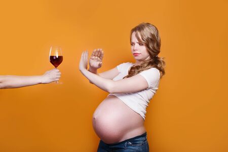 Pregnant woman refuses to drink wine, making stop gesture to glass on yellow backgroundの写真素材