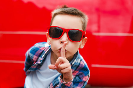 Boy in red sunglasses with finger on lips making a silent gesture in city streetの写真素材