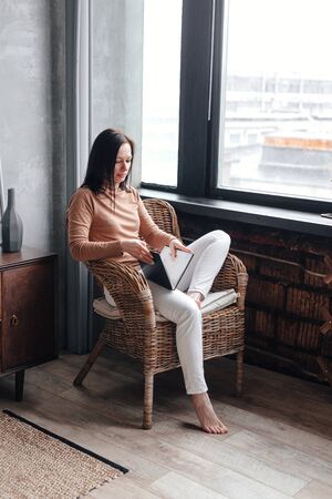 Young woman sitting on chair relaxing in her living room reading book indoorの写真素材