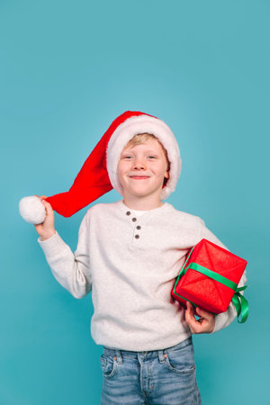funny child in Santa red hat holding Christmas gift in hand on blue background. Christmas concept.