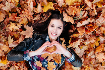 I love autumn. Young smiling woman lying on autumn leaves and making heartの写真素材