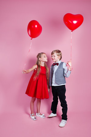 Valentines day. Little boy kissing beautiful little girl on pink background. Children with heart balloonsの写真素材