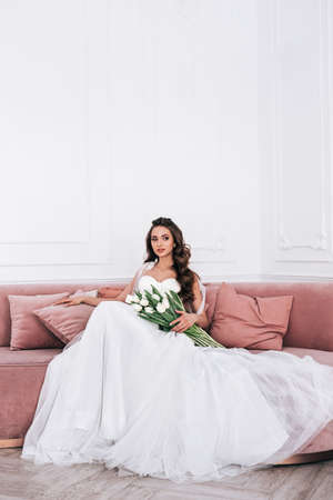 Bride in beautiful dress holding wedding bouquet and sitting on sofa indoorsの写真素材