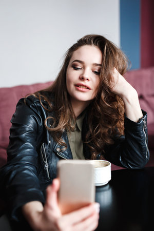 Smiling woman sitting in cafe drinking coffee and using smart phone taking selfie