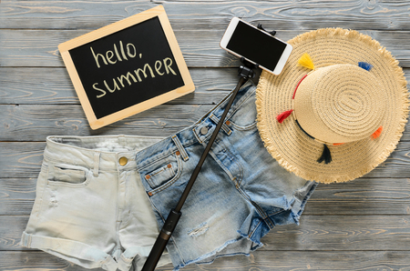 Womens clothing, accessories (denim shorts, straw hat, cellphone, selfie stick, chalkdoard) on grey wooden background. Text Hello summer. Trendy fashion outfit. Shopping, travel concept.  Flat lay. Summer outfitの写真素材