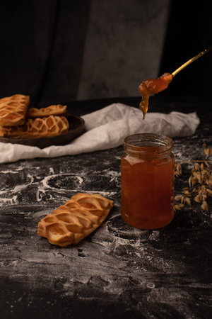 Jam in a glass jar, with a spoon over it and jam buns next to it. Fresh pastries on the table with...