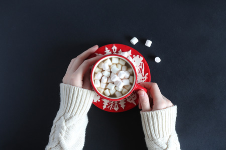 Female hands in white woolen sweater holding red mug with cocoa and marshmallowsの写真素材