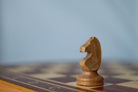 A wooden knight chess piece stands alone on a chessboard against a minimalist soft blue background....