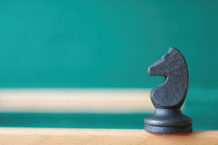 A black wooden knight chess piece stands on a wooden edge against a blurred green background....
