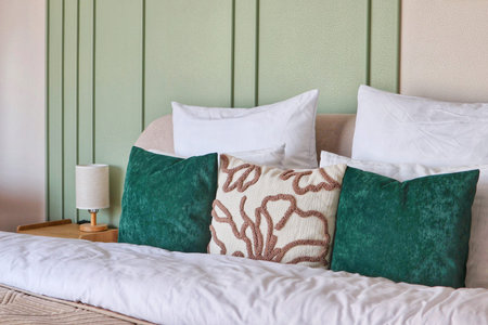 A cozy bed is decorated with white and emerald green pillows against a light green paneled wall in a modern bedroom. Concept of bedroom styling.の写真素材