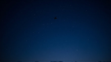Distant airplane cruises through a vast, star-filled night sky above a faint horizonの素材