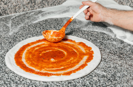 The chef pours tomato sauce over the rolled dough, restaurant concept, kitchen spoon, the process of preparing italian pizzaの写真素材
