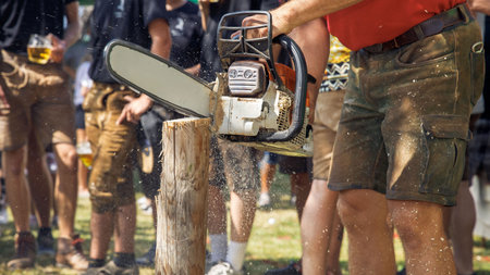 Man using chainsaw to cut wood at outdoor event, surrounded by people, sawdust in air, lively...