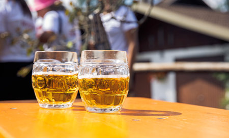 Two clear glass mugs filled with light beer on wooden table, outdoors, sunny day, casual gathering, refreshment, relaxation, beer mugs conceptの写真素材