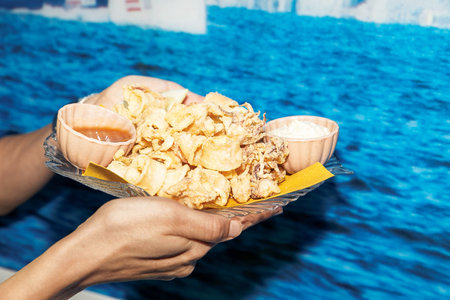 Platter of crispy fried calamari, lemon, dipping sauces served, hands holding plate, sea background, delicious seafood conceptの写真素材
