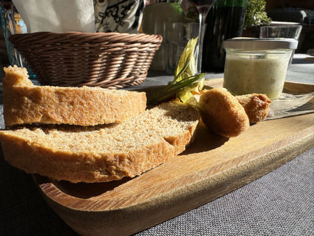 Fresh bread slices with fish croquette and sauce on wooden platter in sunlight.