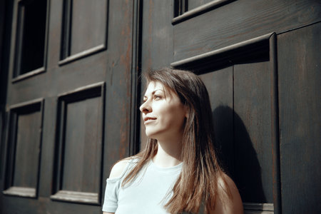 A young woman looking thoughtfully to the side, leaning against a dark wooden door, softly lit by the sunの写真素材