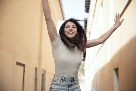 A cheerful young woman jumping energetically, arms raised, in a bright sunny alleyway, full of lifeの写真素材