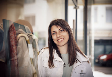 A cheerful young woman in a white jacket smiling brightly while standing near a shop window in the cityの写真素材