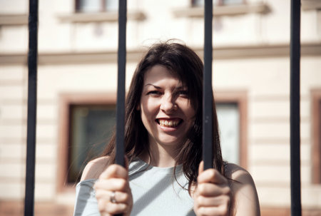 A cheerful young woman standing behind black bars, smiling brightly in an urban outdoor settingの写真素材