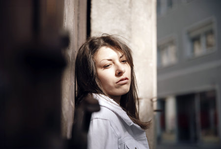 A young woman gazing pensively while leaning on a wall, illuminated by soft sunlight in a cityの写真素材