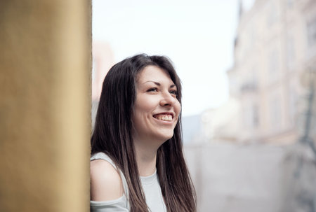 A cheerful young woman smiling brightly, leaning against a wall with urban architecture in the backgroundの写真素材