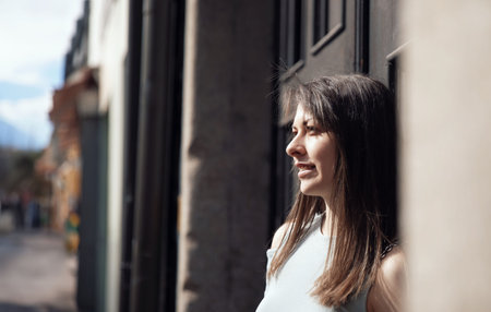A young woman smiling gently while leaning against a wall in a sunny urban street, looking peacefulの写真素材